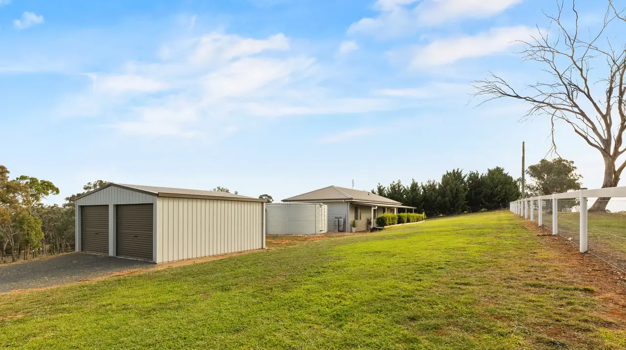 Colorbond shed with two roller doors on a gravel pad, cottage beyond on the lawn